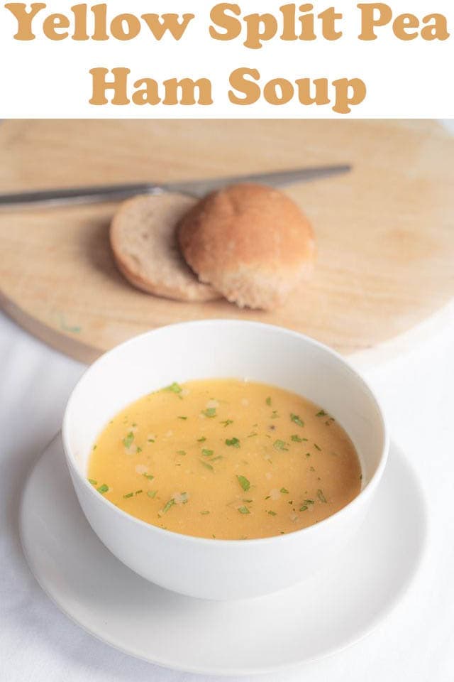 A bowl of split pea soup with a roll halved on a chopping board in the background.