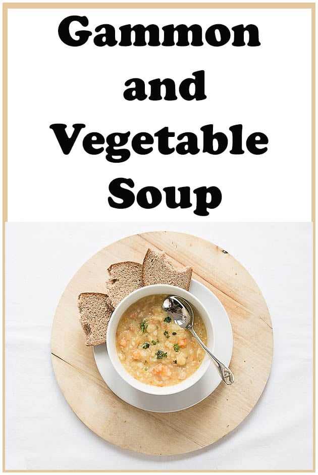 A bowl of ham and vegetable soup on a board with a spoon in it. Slices of bread around the board.