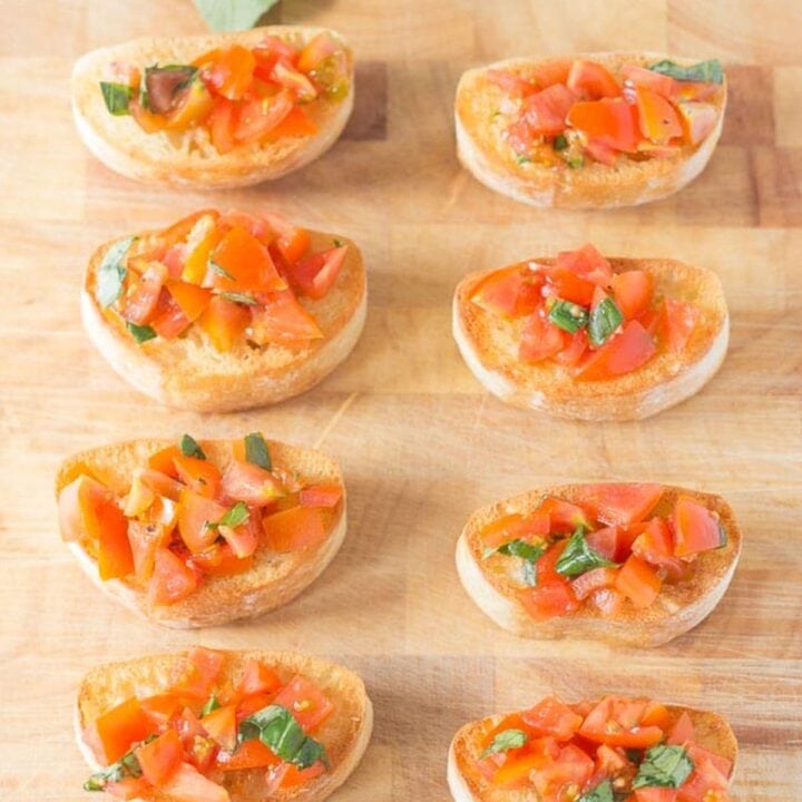 A bread board with 10 slices of mini bruschetta with cherry tomatoes and basil on.