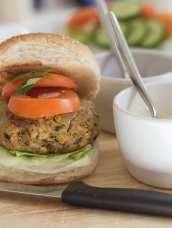 A chickpea burger in a burger bun with sliced tomotoes on top. A small bowl of mayonnaise with a spoon in to the right.