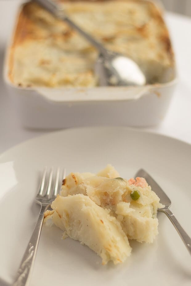 A portion of fish pie served on a plate with the rest of the pie in the background.