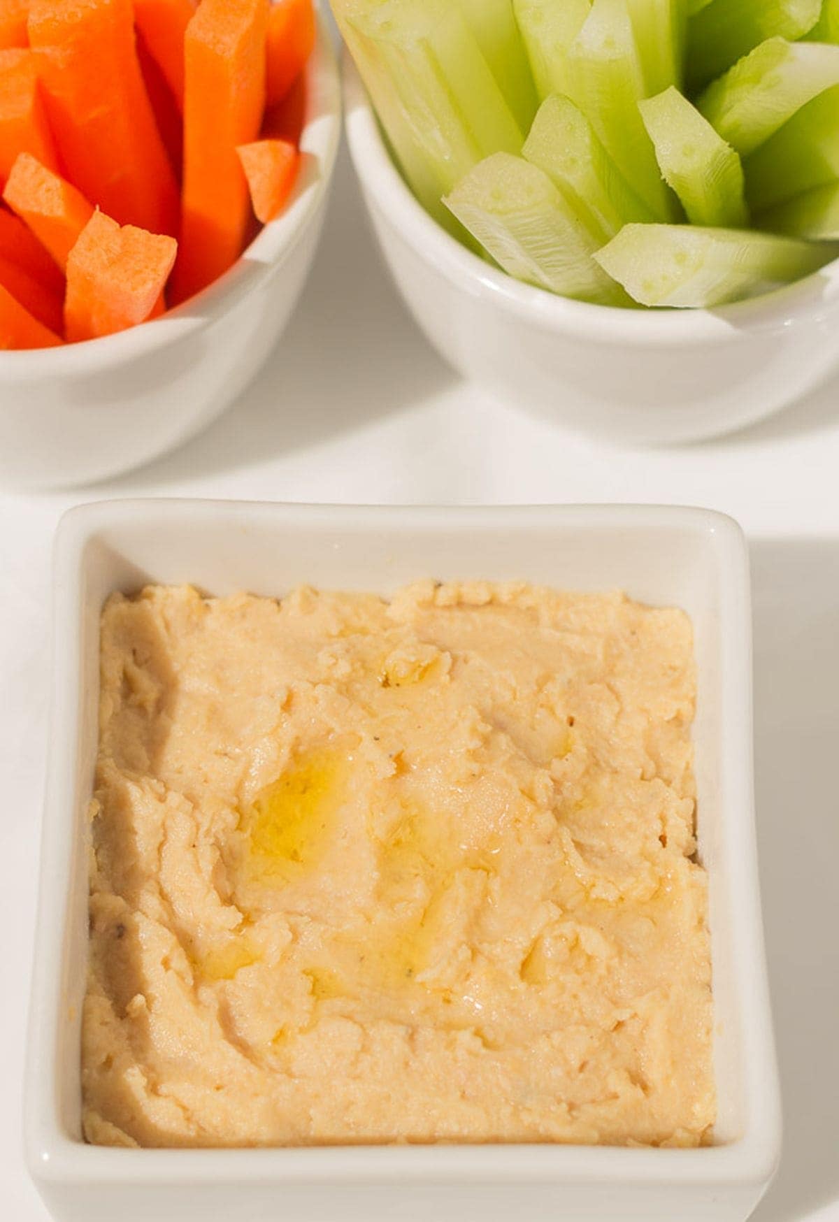 A white dish of low fat hummus dips with baton carrots and celery in the background.