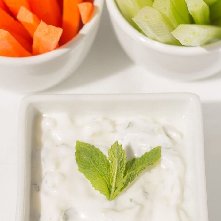 Low fat tzatziki dip in a small square serving dish with a sprig of mint leaves on top and crudities in the background.