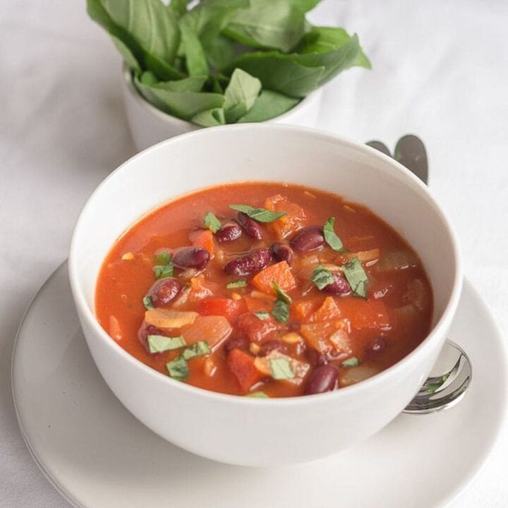 A bowl of red pepper tomato and kidney bean soup with a dish of basil leaves in the background.