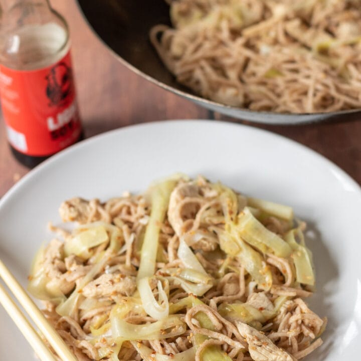 A plate of stir fried chicken with leek and noodles. Chop sticks to the side and a bottle of soy sauce and wok above.