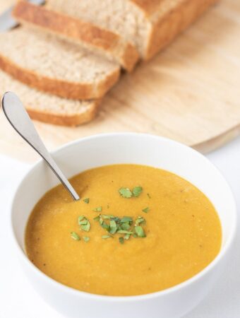 A bowl of carrot and lentil soup, garnish and with a spoon in it. A loaf of sliced bread on a breadboard in the background.