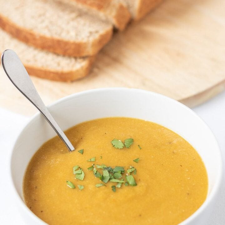 A bowl of carrot and lentil soup, garnish and with a spoon in it. A loaf of sliced bread on a breadboard in the background.