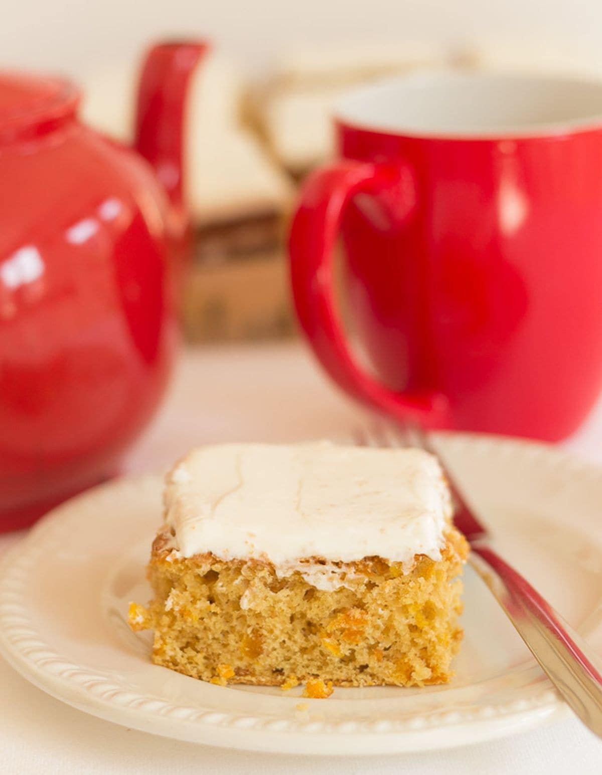 A slice of orange and apricot tray bake on a white plate with a pot of tea and cup in the background.