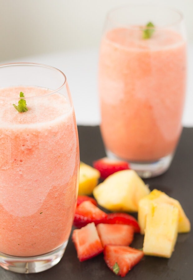 Two glasses of strawberry sunrise smoothie. Diagonally in front of the other with sliced pineapple and strawberries in between.