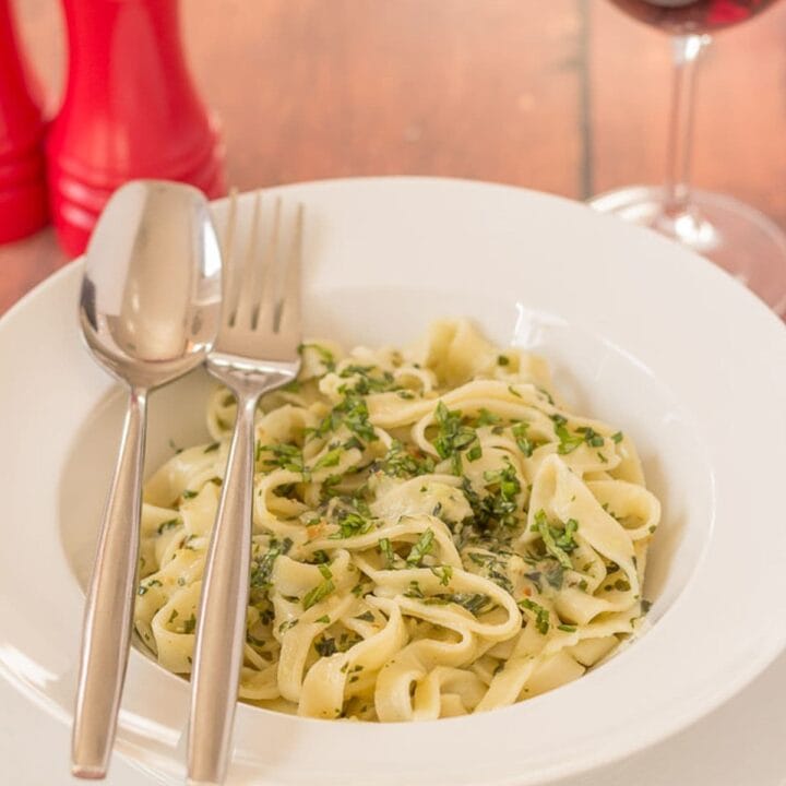 A bowl of homemade tagliatelle and pesto knife and fork to the side and a glass of red wine and salt and pepper shakers in the background.