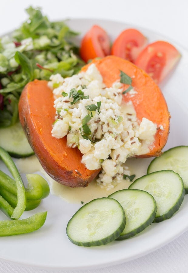 Close up of a baked sweet potato topped with feta and orange on a plate. A salad of cucumber, lettuce and tomatoes surrounding.