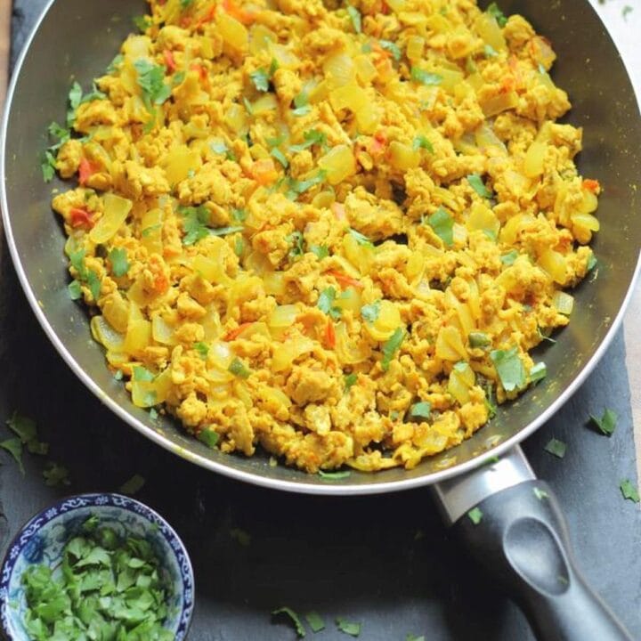 Birds eye view of a large pan of Indian scrambled eggs on a black slate with a dish of chopped coriander to the bottom left.