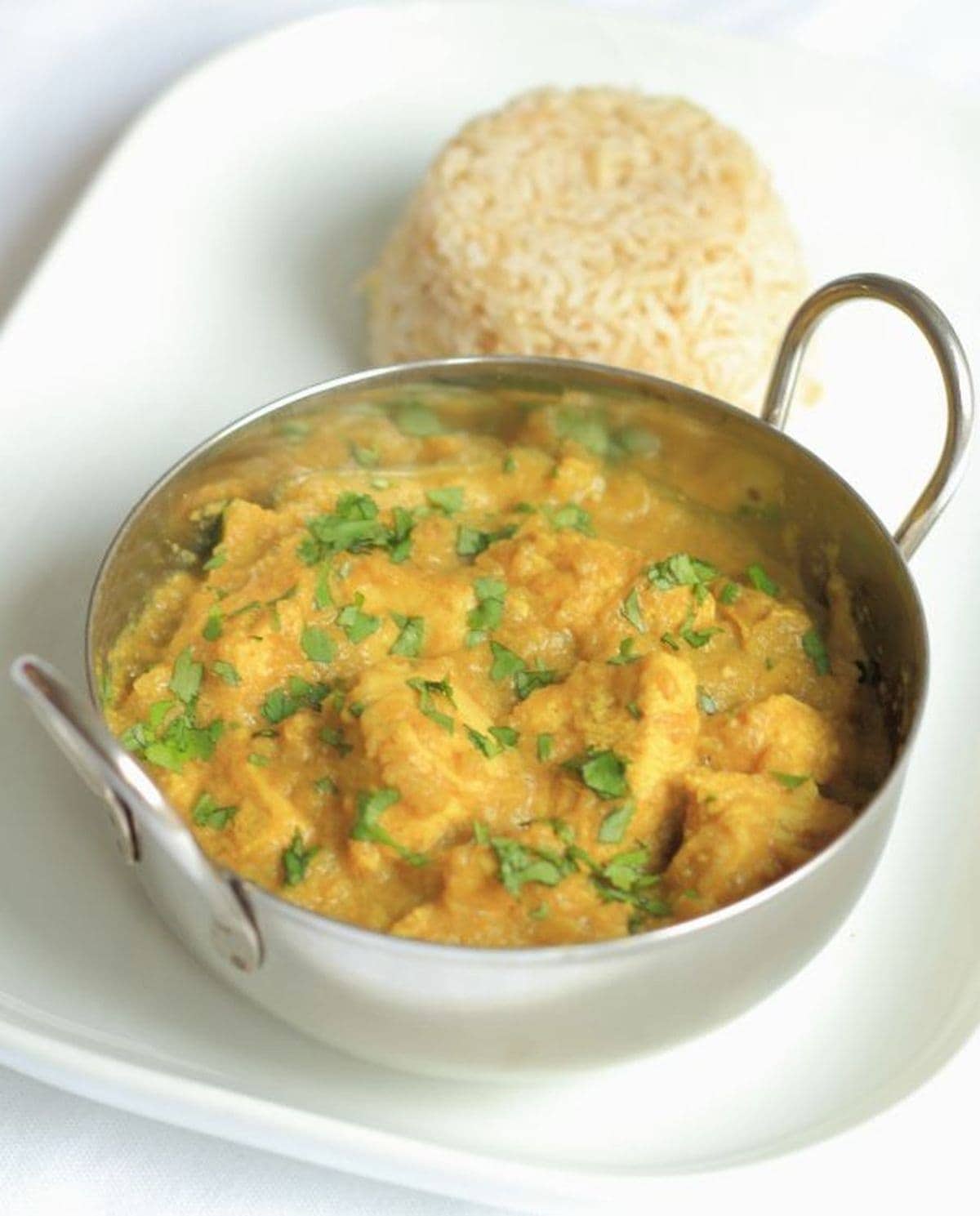 A balti dish of lighter chicken korma on a serving plate with a portion of wholegrain rice in the background.