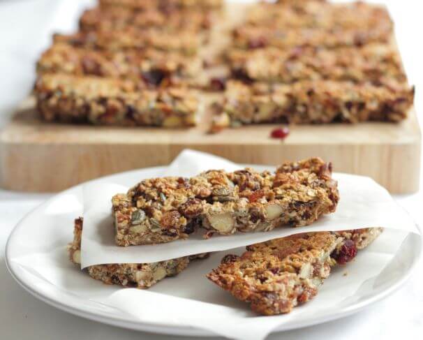 Close up of three fruit and nut snack bars on a plate separated by greasproof paper. Chopping board with the rest of the bars on in the background.