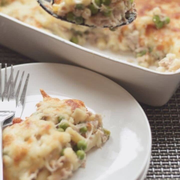 A portion of tuna macaroni bake being lifted from the baking dish. A portion sitting on a plate at the front with 2 forks on.