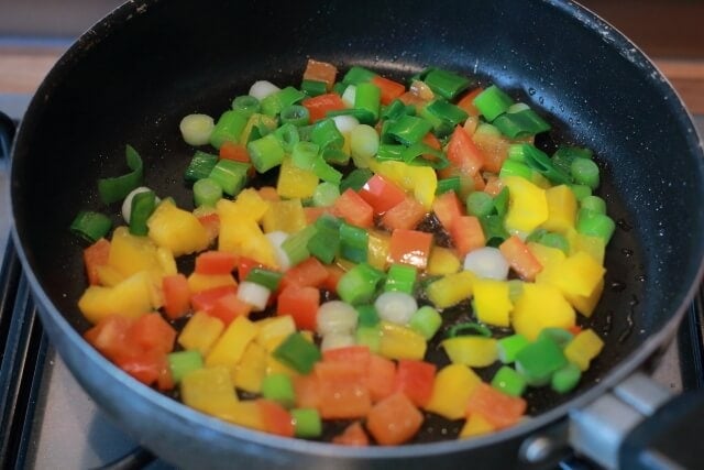 A poaching pan of diced yellow and orange peppers and chopped spring onions cooking.