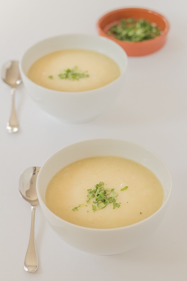 Two bowls of potato and onion soup with spoons to the left hand side and a dish of chopped parsley in the background.