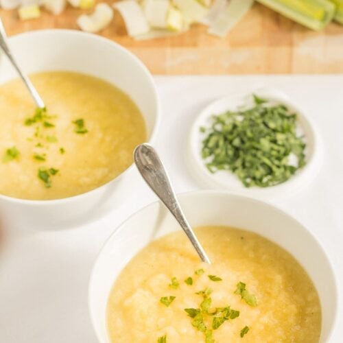 Two bowls of Scottish leek and potato soup diagonally across from each other with spoons in. Assortment of chopped vegetables around the top of the bowls.