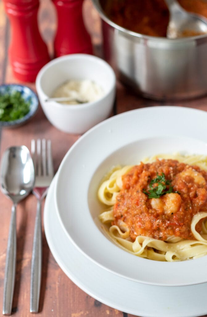A bowl of tagliatelle prawns in tomato and pepper sauce. Fork and spoon to the left side. Pan of sauce and dishes of grated parmesan and chopped parsley above.