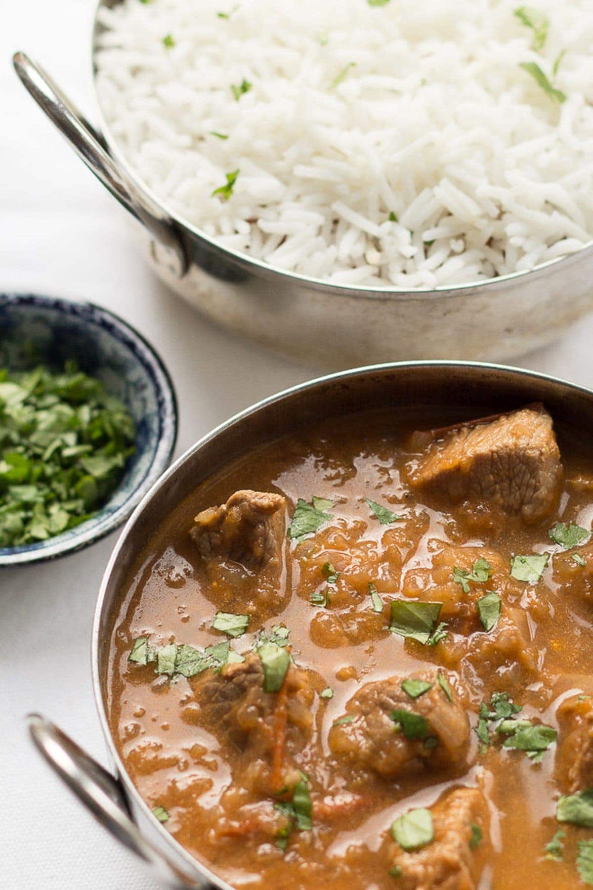 A balti dish of skinny lamb curry with a small dish of chopped coriander and a balti dish of rice in the background.
