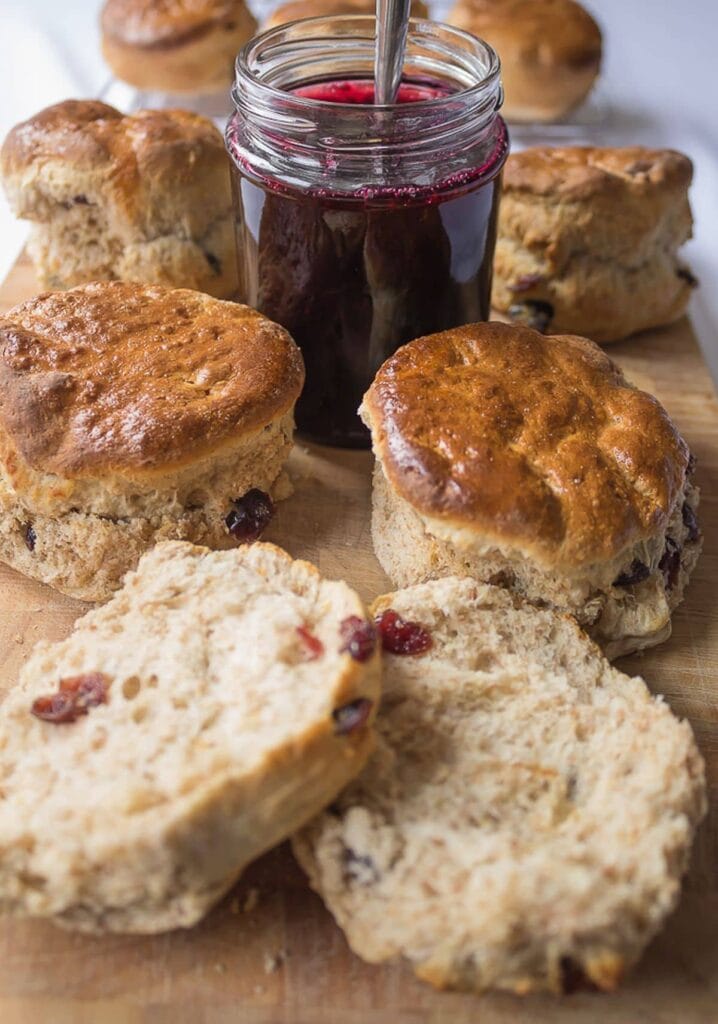 A chopping board with wholemeal cranberry scones on and a pot of jam with a spoon in the middle. A scone cut in half at the front.