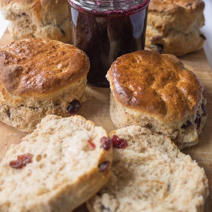 A chopping board with wholemeal cranberry scones on and a pot of jam with a spoon in the middle. A scone cut in half at the front.