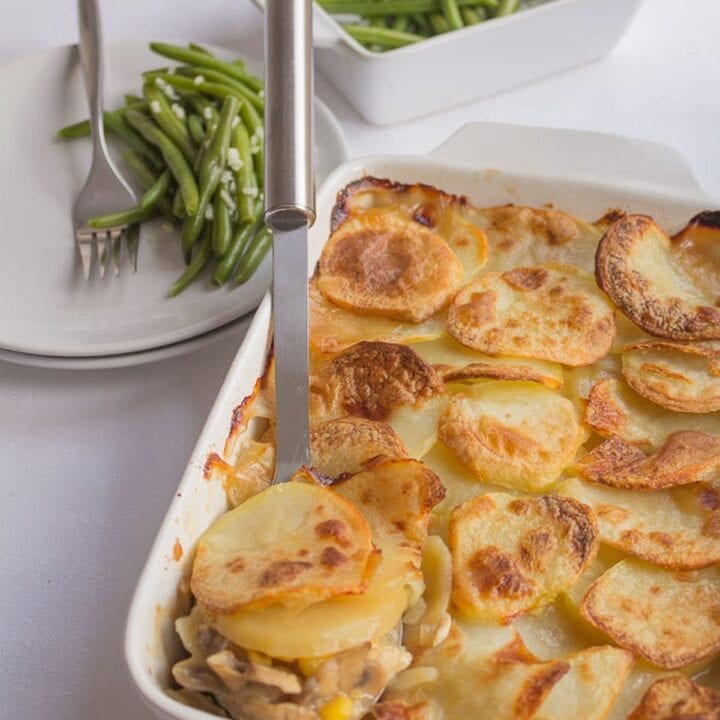 A casserole dish of chicken sweetcorn and mushroom bake with a spoon lifting out a portion. Green beans in a serving dish and on a dinner plate in the background.