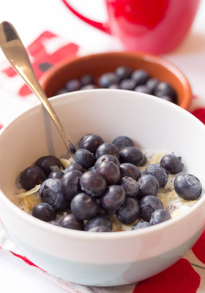 Close up of a bowl of porridge with blueberries and almonds with a spoon in. A dish of blueberries and coffee cup in the background.