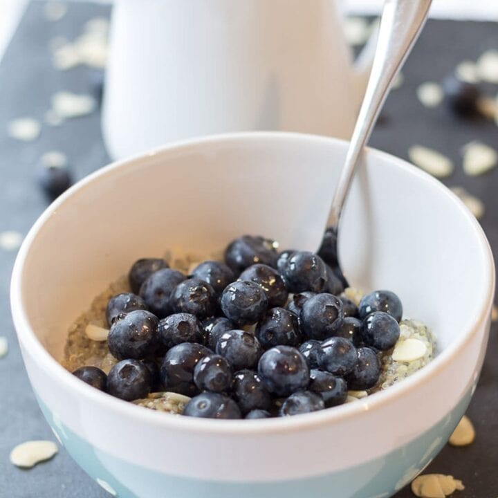 A bowl of vanilla quinoa blueberry breakfast with a spoon in and a milk jug in the background.