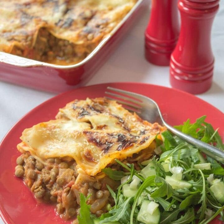 A portion of green lentil lasagne served with side salad on a plate. The rest of the lasagne dish and salt and pepper cellars in the background.