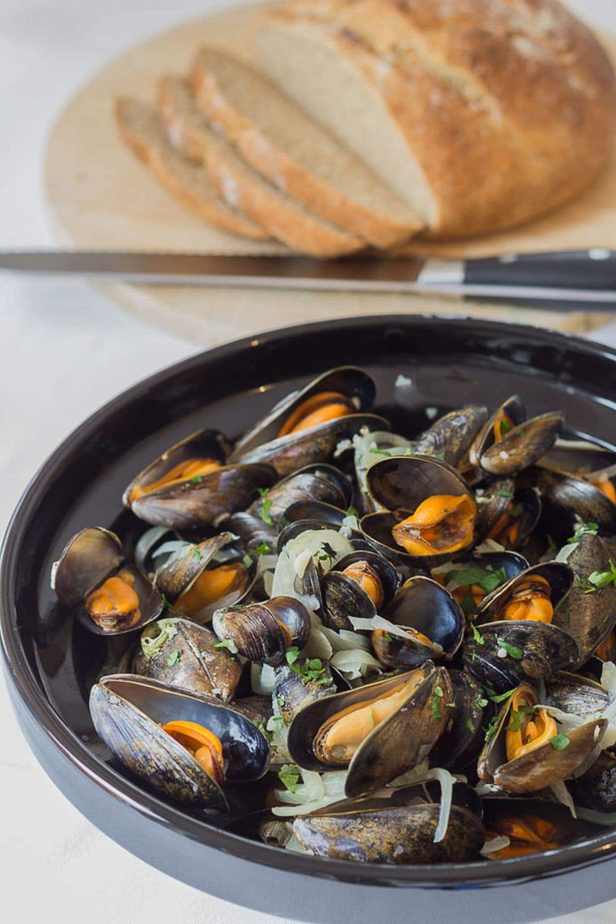 Cooked moules marinieres in a large bowl with a freshly made sliced cob loaf in the background.