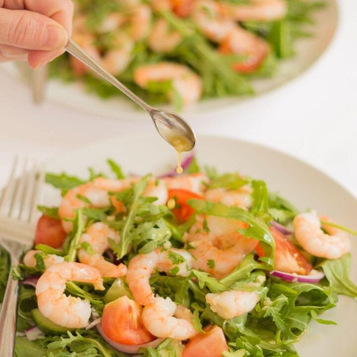 Two plates of king prawn and rocket salad one in front of the other. Dressing being added by teaspoon to the front plate.
