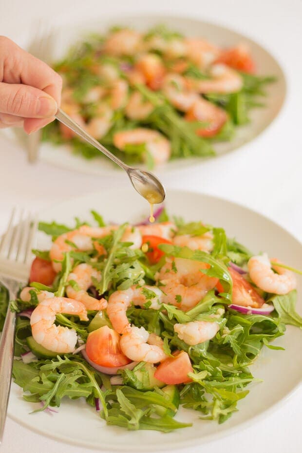 A teaspoon of lime dressing being dropped over a plate of king prawn and rocket salad. Another plate of the salad behind.