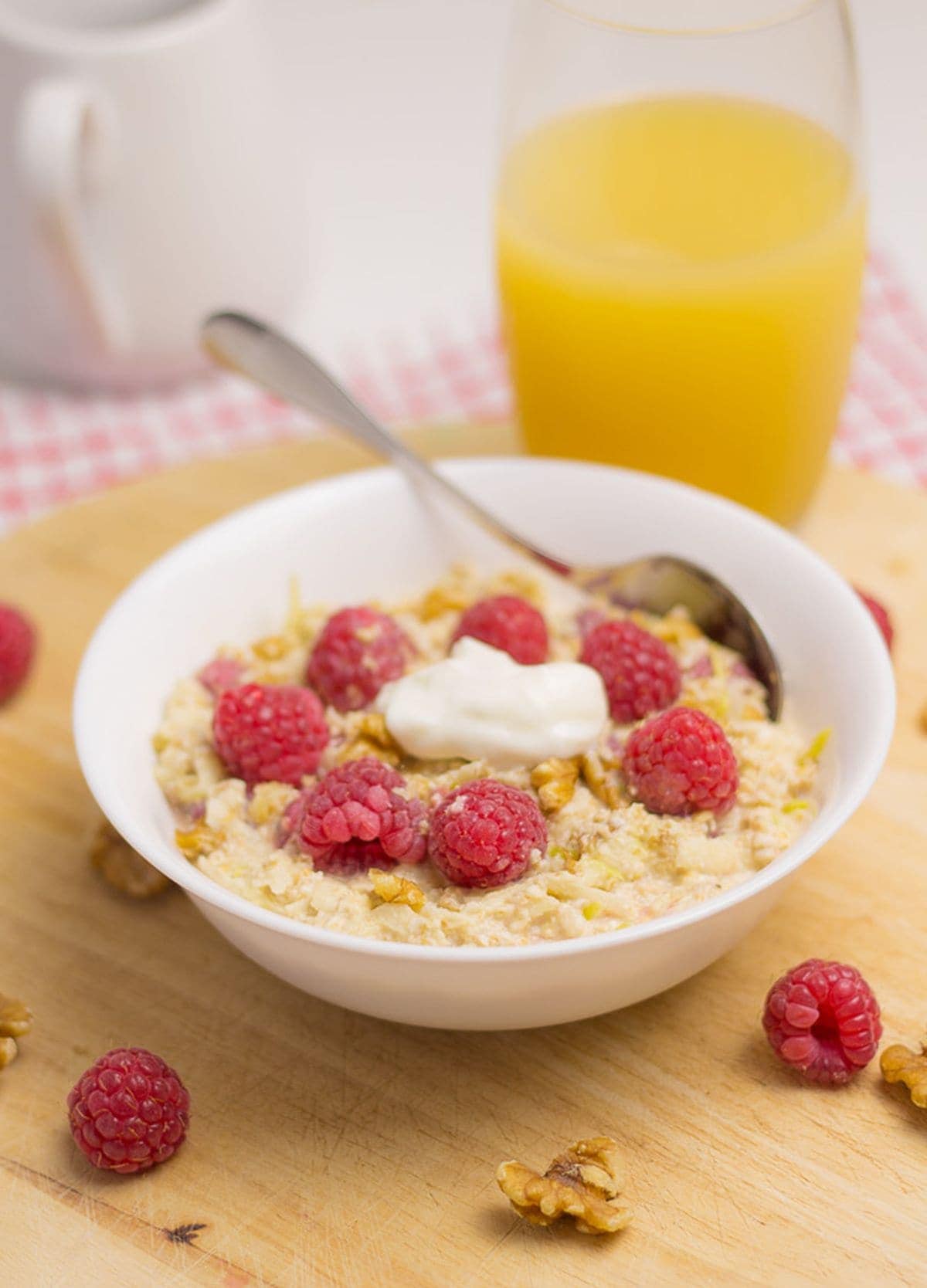 A bowl of raspberry bircher overnight oats topped with yogurt and extra raspberries. A glass of orange juice in the background.
