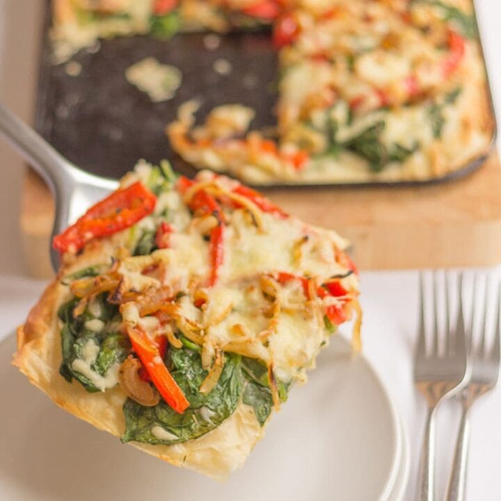 A slice of red pepper and spinach filo tart being lifted onto a plate by a slce from the red of the tart on the tray in the background.