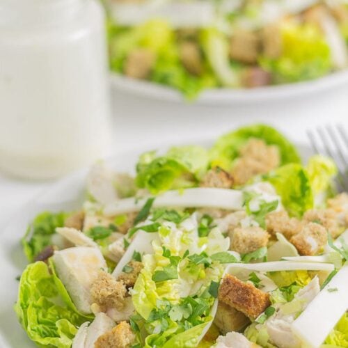 Two plates of chicken caesar salad one in front of the other with a jar of dressing in between.