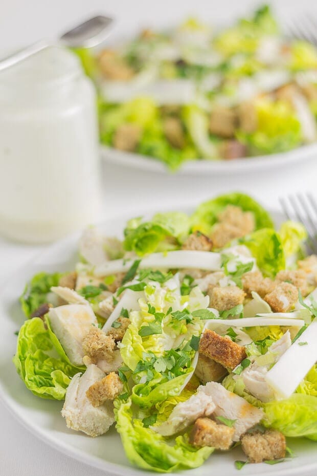 A plate of chicken Caesar salad with a jar of dressing behind and then another plate of similar salad.