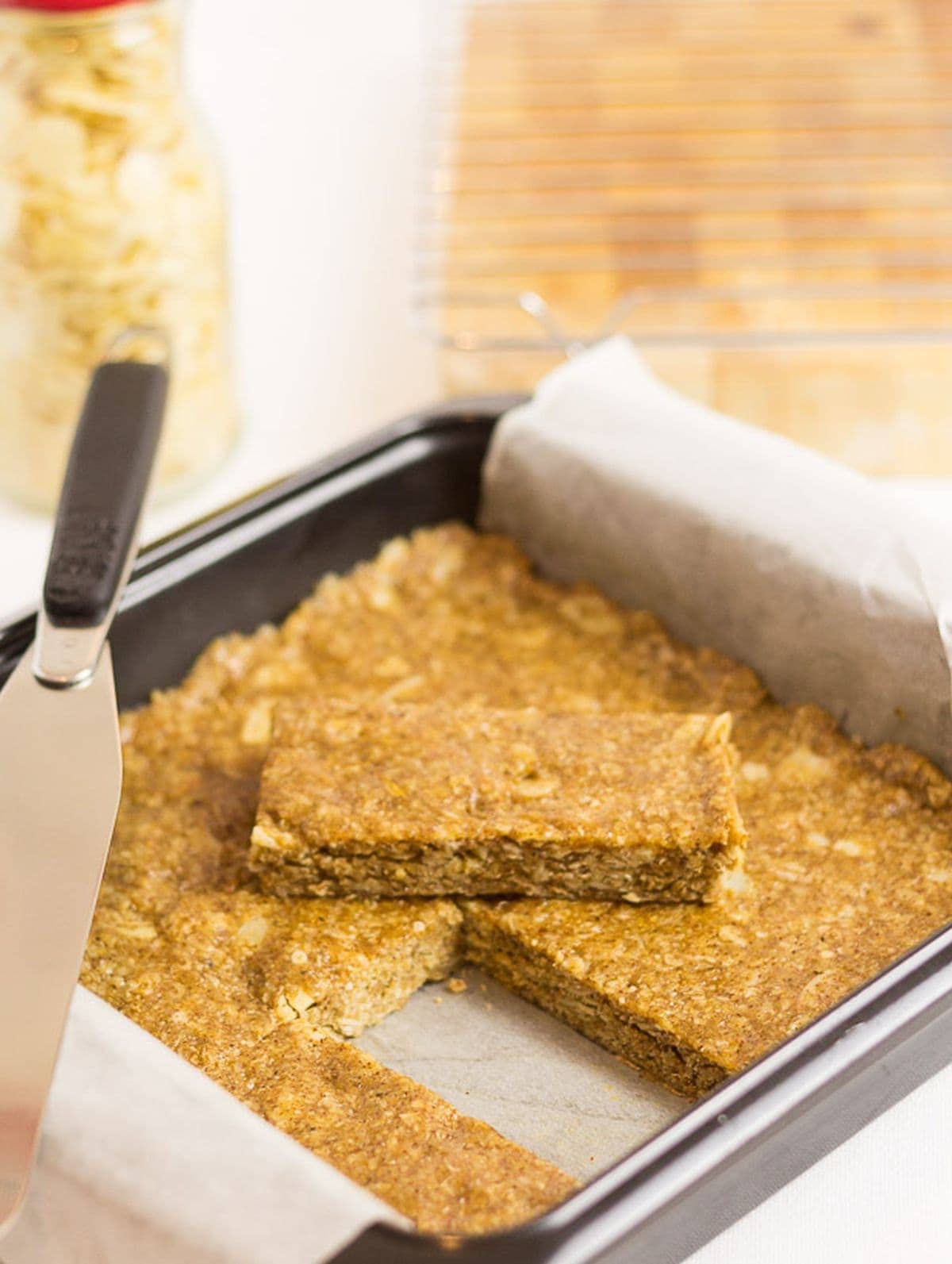 A square tin of no-bake almond oat bars. One bar removed and placed on top with a spatula to the side.