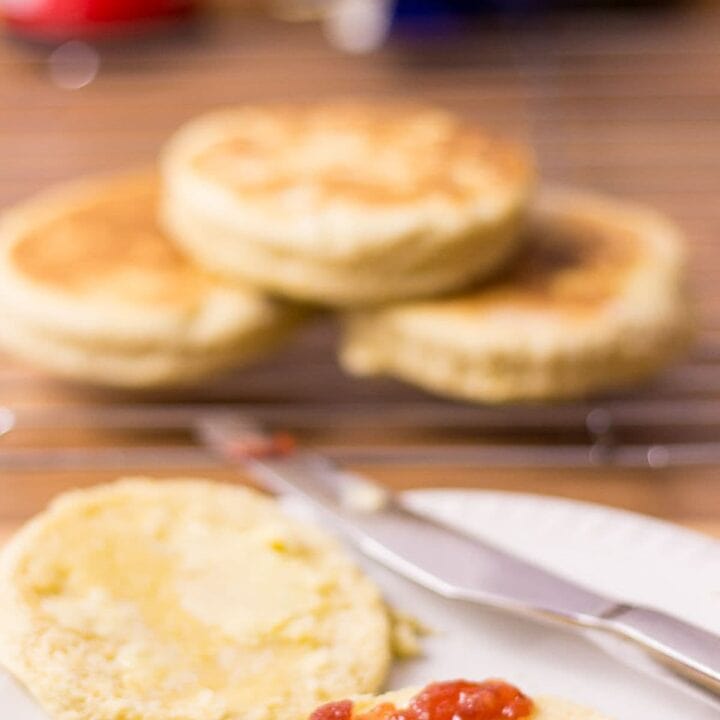 Two quick English muffins on a plate. One with jam on and a bite taken out. The rest of the muffins on a wire rack in the background.