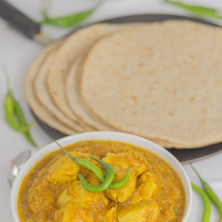 A bowl of green chilli chicken curry garnished with green chillies and some paratha breads on a tava in the background.