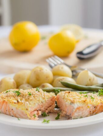 A plate with oven baked salmon with cream cheese served with baby potatoes and green beans. A chopping board with lemons on in the background.