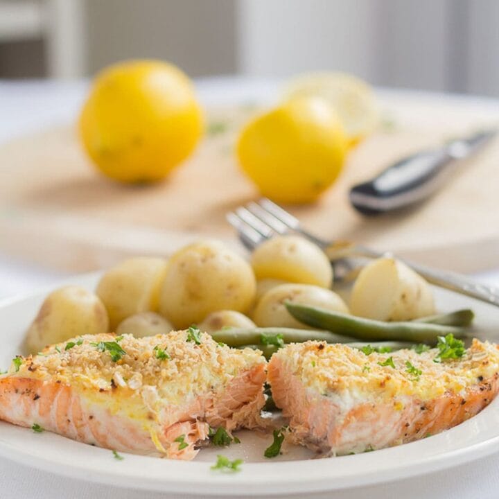A plate with oven baked salmon with cream cheese served with baby potatoes and green beans. A chopping board with lemons on in the background.