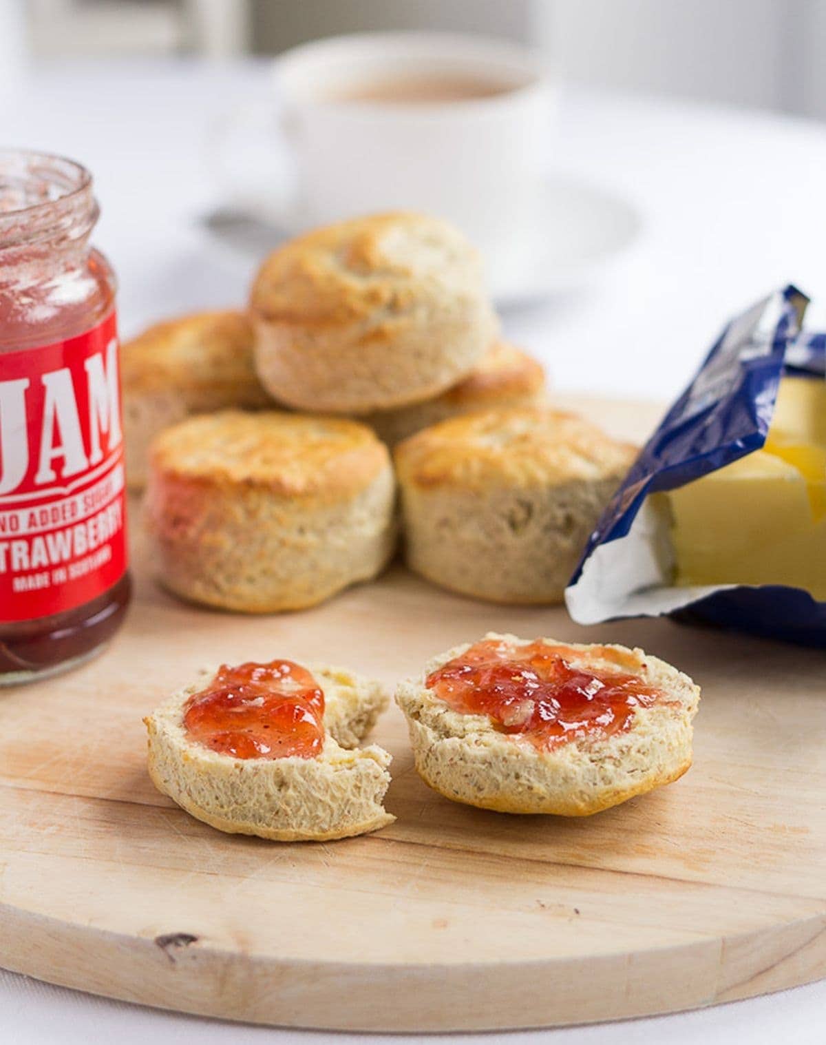 A Scottish bran scone cut in half and covered with butter and jam on a chopping board. More scones in the background as well as a jar of jam, block of butter and a cup of tea.