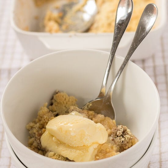 Close up of a bowl of sugar free cinnamon apple crumble and ice cream with two serving spoons in.