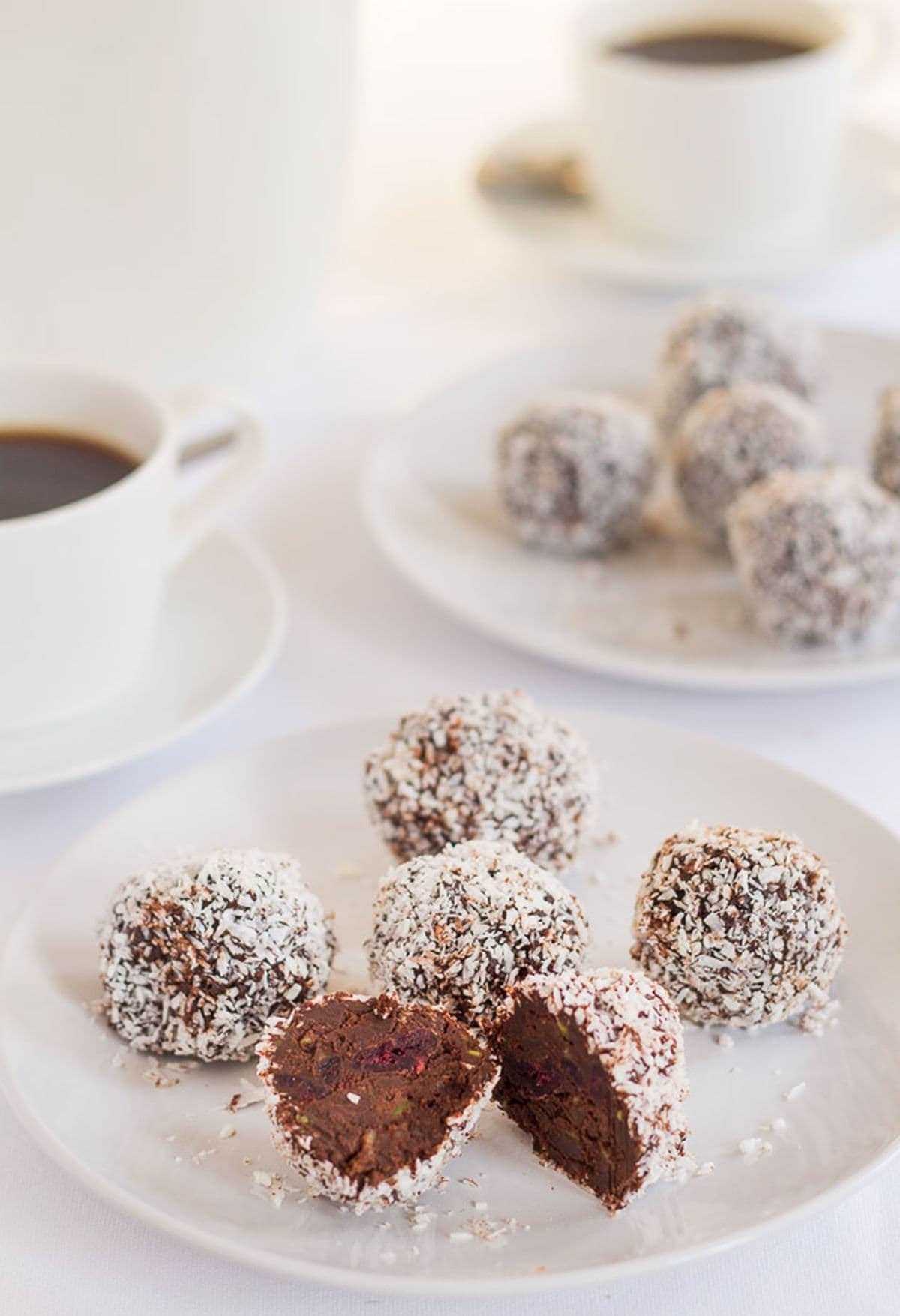 A plate of chocolate cranberry truffles with one cut in half. Another plate in the background as well as two cups of coffee.