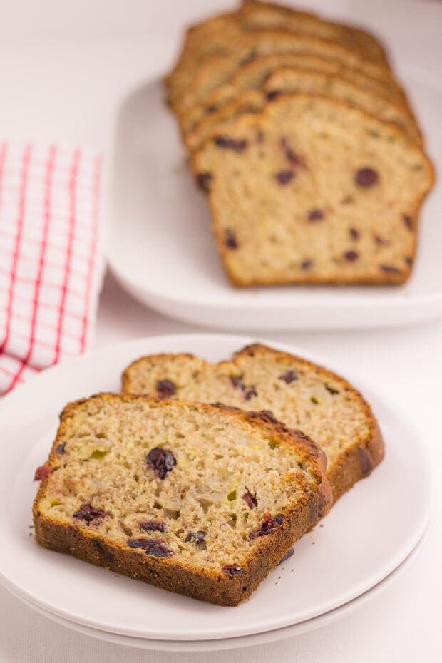 Two slices of cranberry banana bread on a plate. The rest of the cake on a serving dish in the background.