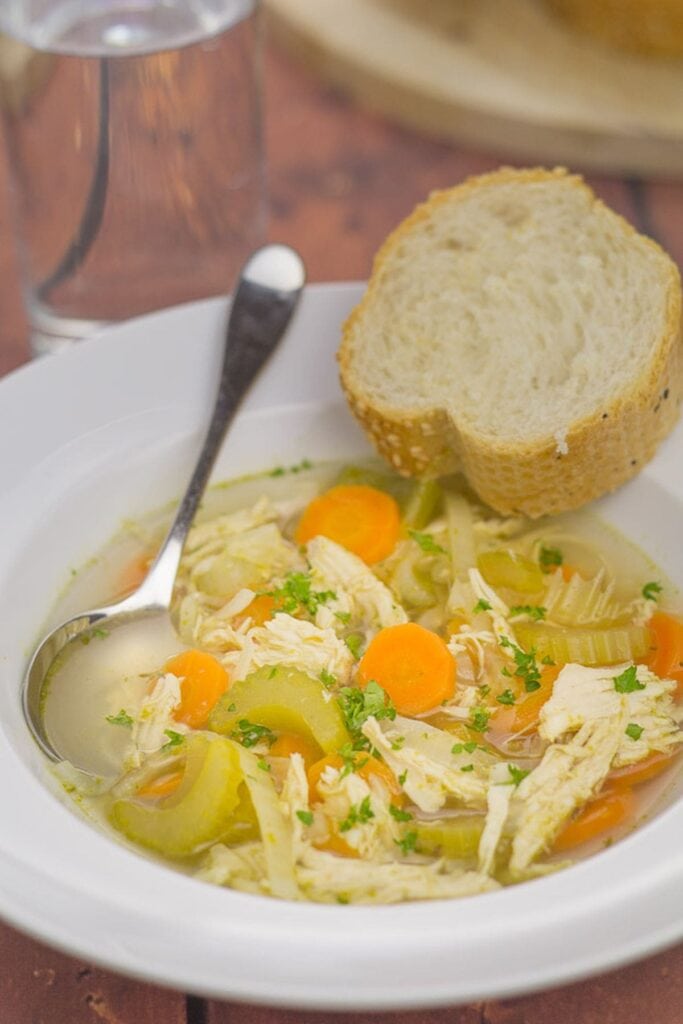A bowl of simple chicken and vegetable soup with a spoon in. Served with a slice of bread on the side.