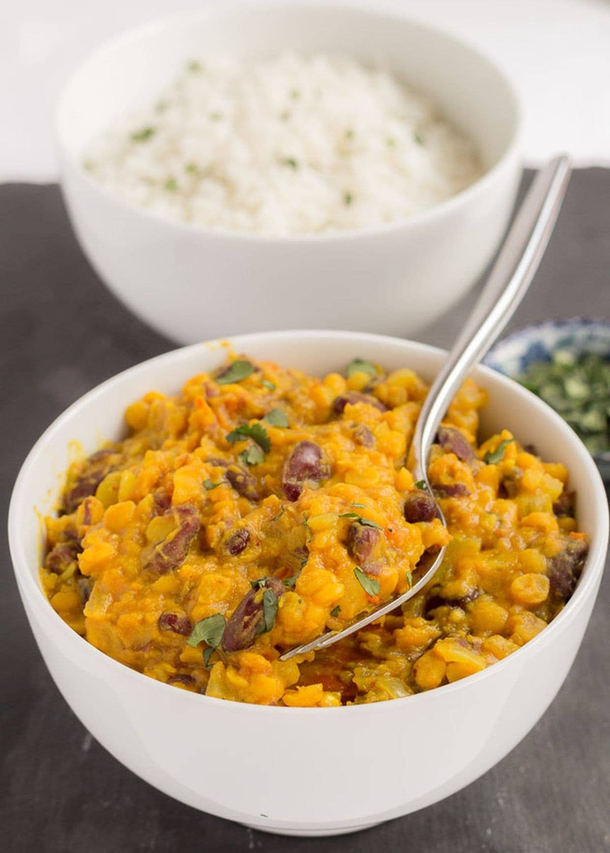 A bowl of lentil and kidney bean dal served with a spoon in it. A bowl of rice and dish of coriander to garnish in the background.