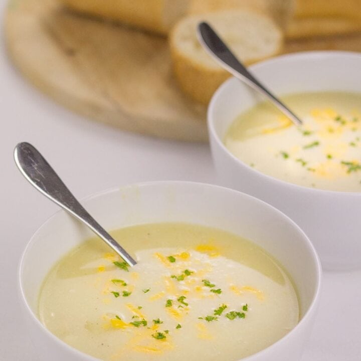 Two bowls of cheesy cauliflower and white bean soup with spoons in. One in front of the other a bread board with sliced baguette on in the background.