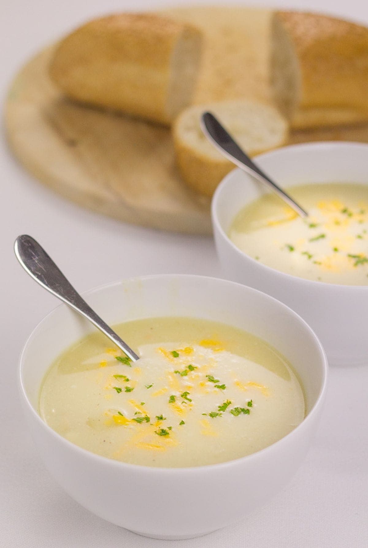 Two bowls of cheesy cauliflower and white bean soup with spoons in. One in front of the other a bread board with sliced baguette on in the background.